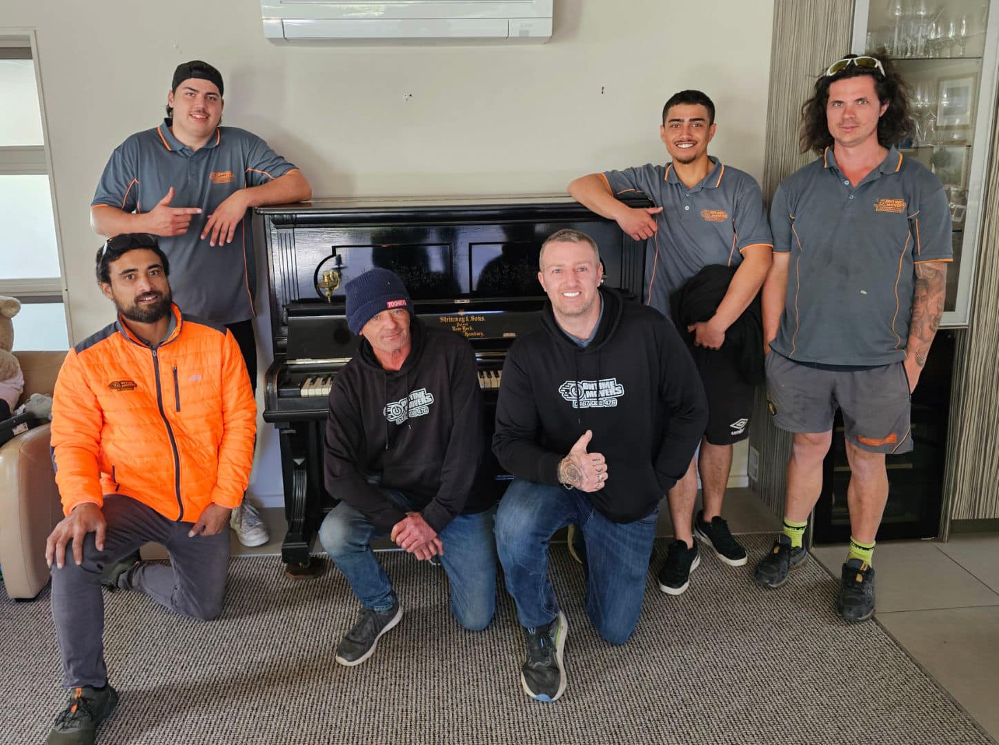 Ontime Movers team posing indoors in front of a black upright piano, wearing branded uniforms and casual workwear